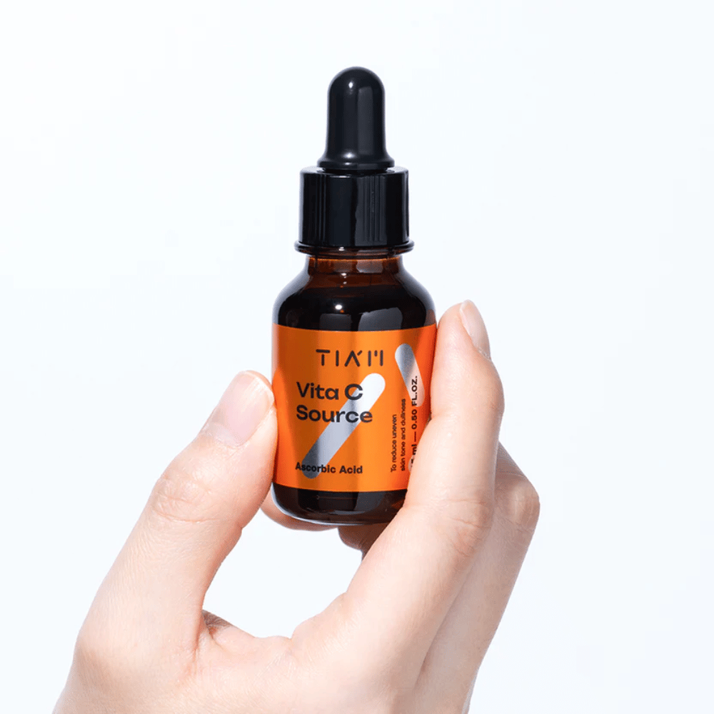 Ręka trzyma TIA'M Vita C Source, serum z witaminą C w brązowej szklanej butelce z pipetą. Produkt jest zaprojektowany, aby wyrównywać koloryt skóry i nadawać jej blask.