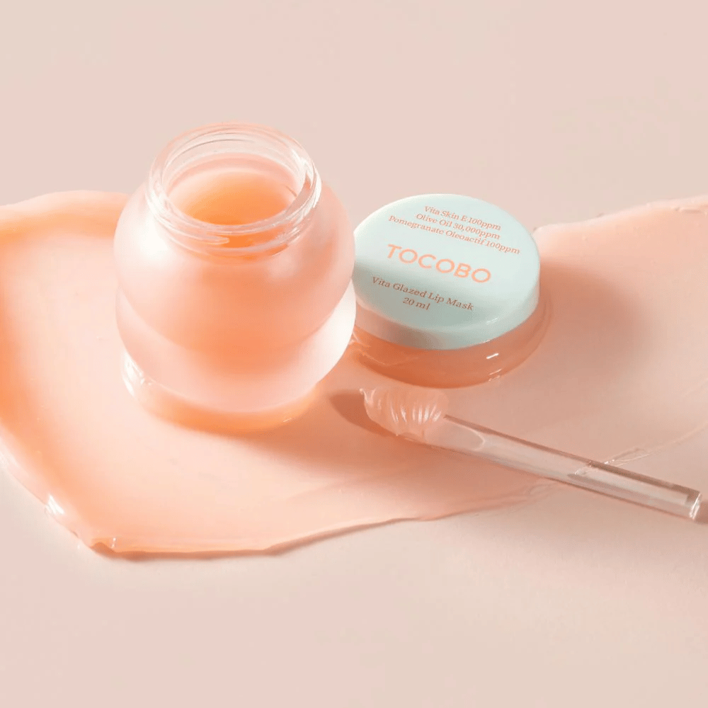 Pojemnik z "Vita Glazed Lip Mask" od Tocobo jest pokazany. Pojemnik jest przezroczysty z jasnoturkusową pokrywką i zawiera błyszczącą, różową maskę do ust. Obok pojemnika leży mała szpatułka, a część maski jest rozprowadzona na podłożu. Produkt jest zaprojektowany do nawilżania i pielęgnacji ust.