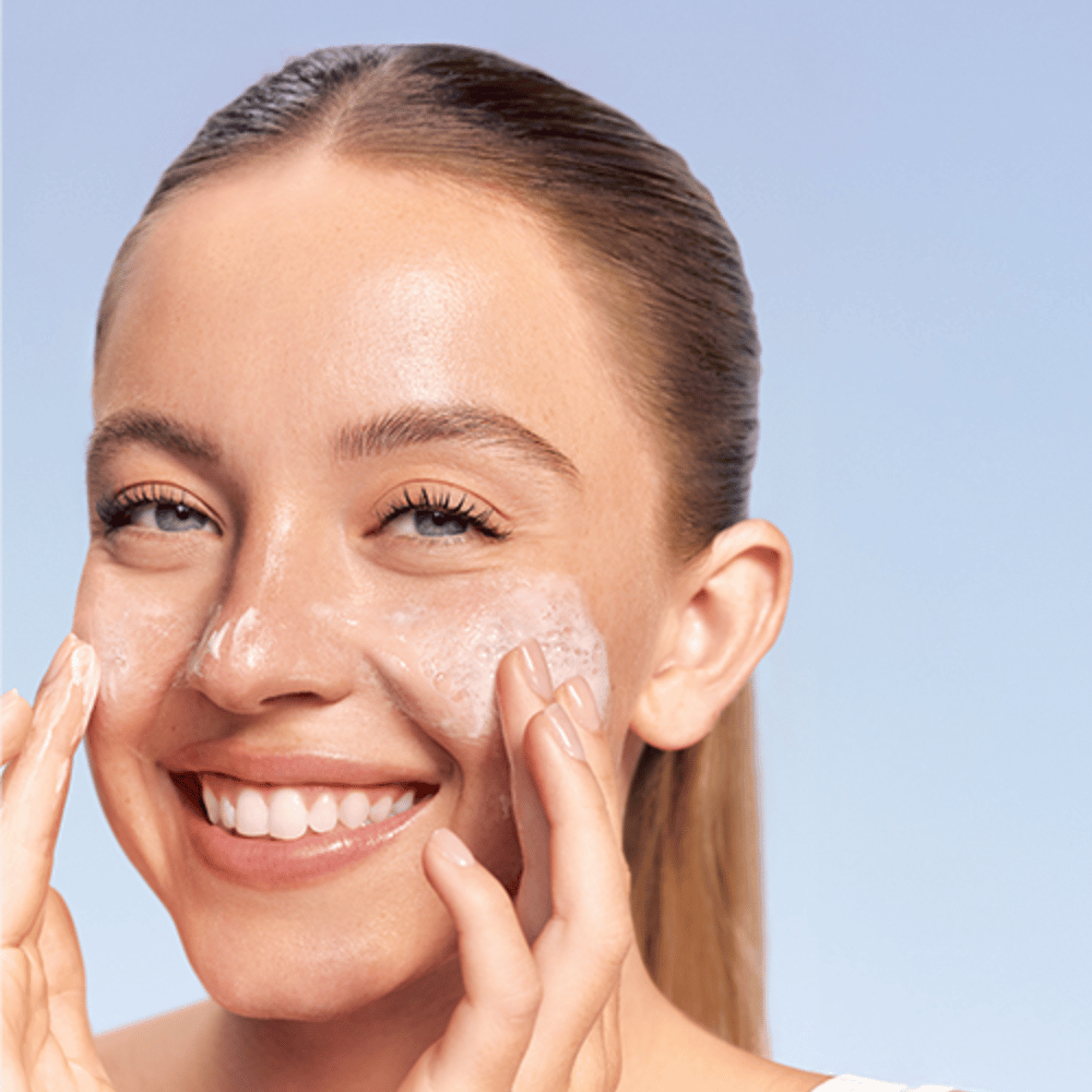 Uśmiechnięta kobieta nakłada LANEIGE Water Bank Gentle Gel Cleanser w formie piany na policzki, na niebieskim tle.