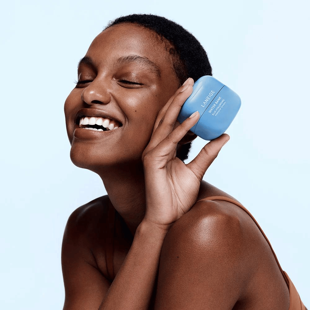 Uśmiechnięta kobieta trzyma niebieski słoik LANEIGE Water Bank Blue Hyaluronic Cream przy policzku, na jasnoniebieskim tle.