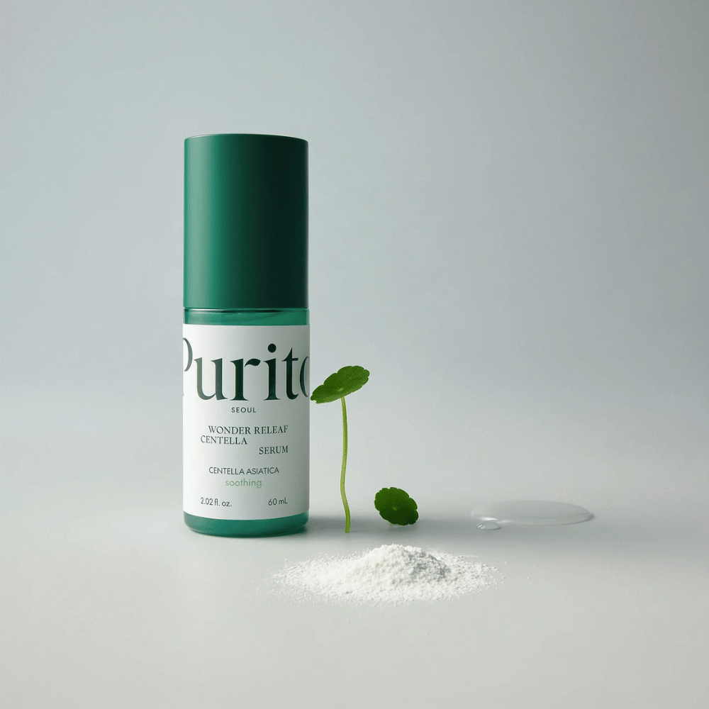 Grön flaska av Purito Wonder Releaf Centella Serum 60 ml står på ett vitt underlag, omgiven av centellablad, ett genomskinligt serumdropp och ett litet vitt pulver.