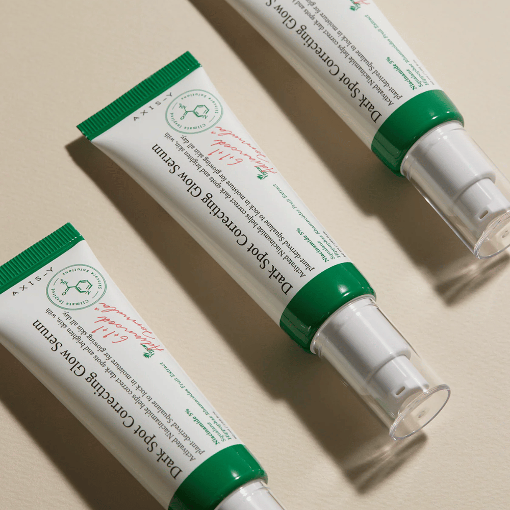 Trzy tuby Axis-Y Dark Spot Correcting Glow Serum z zieloną nakrętką i białym opakowaniem leżą na jasnym podłożu.