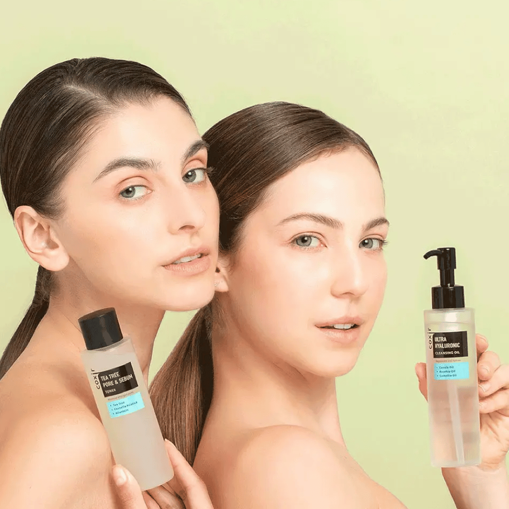 Dwie kobiety z odkrytymi ramionami pozują z produktami do pielęgnacji skóry; jedna trzyma butelkę oznaczoną "TEA TREE PORE & SEBUM", a druga butelkę z pompką oznaczoną "ULTRA HYALURONIC CLEANSING OIL". Stoją na tle jasnzielonym.