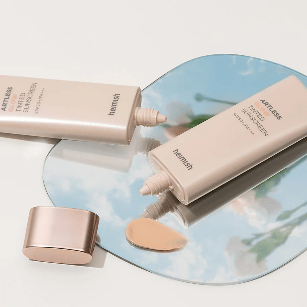 Butelka heimish Artless Glow Tinted Sunscreen SPF50+ PA+++ 40ml i butelka concealer na lustrze, które pokazuje różne odcienie skóry.